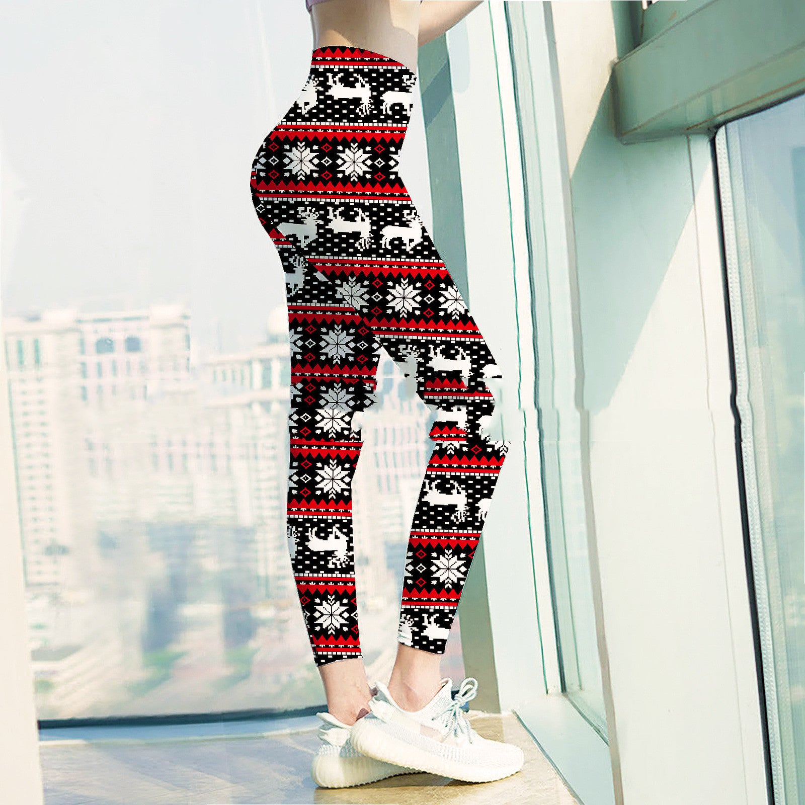Woman wearing festive leggings with reindeer and snowflakes, standing by a large window. The leggings are black, red, and white. She is wearing white sneakers.
