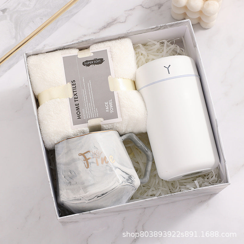 Gift box containing a fluffy white face towel, a sleek white insulated tumbler, and a marble-patterned mug labeled “Fine.” The box has white shredded paper lining.