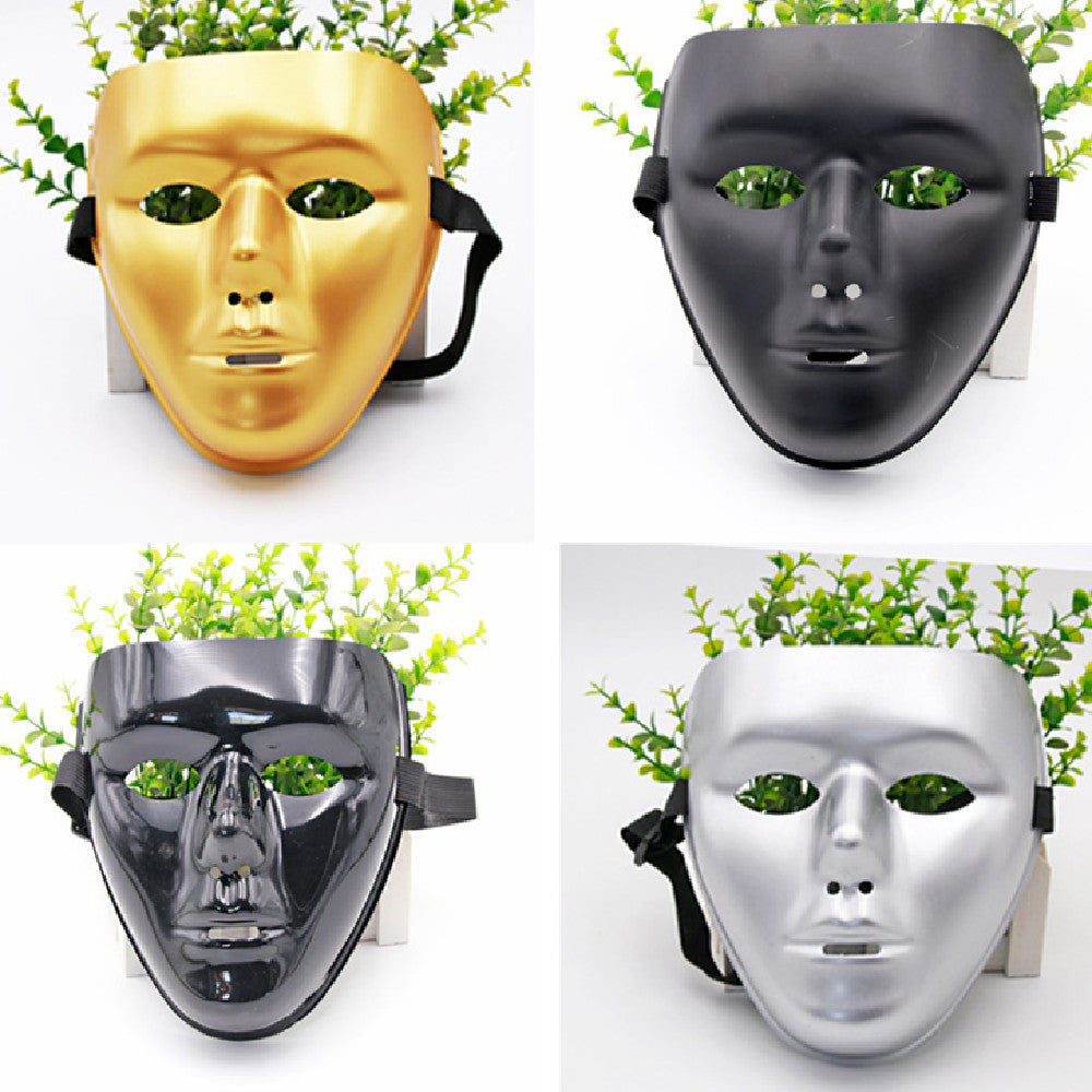 Four face masks are displayed: gold, matte black, glossy black, and silver. Each mask has eye and mouth openings, with green plants arranged behind them for a decorative effect.