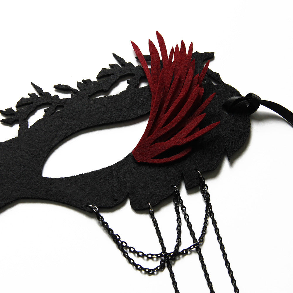 Decorative black masquerade mask with intricate cut-out design, featuring bold red feather details and dangling black chains, set against a white background.