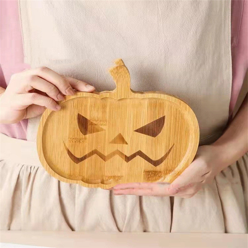 Person holding a wooden pumpkin-shaped cutting board with a carved-out jack-o'-lantern face, against a light-colored apron.