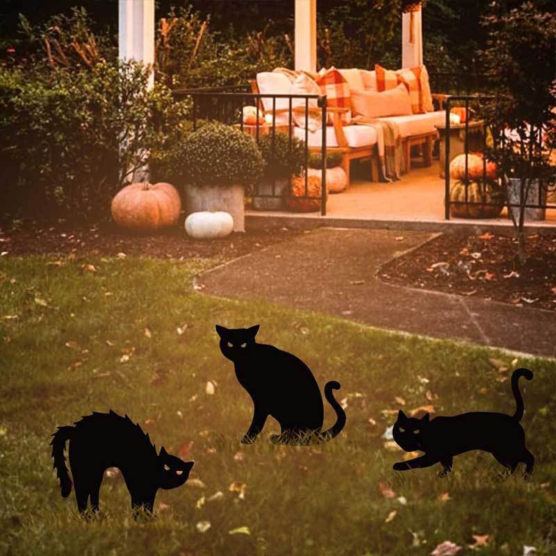 Three black cat silhouettes on a lawn near a cozy patio decorated with pumpkins and cushions. The warm, inviting setting suggests a festive autumn atmosphere.