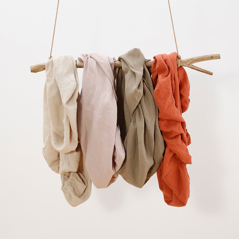 Four textured fabric pieces in beige, pink, olive, and rust hang on a wooden branch suspended by ropes against a white background.