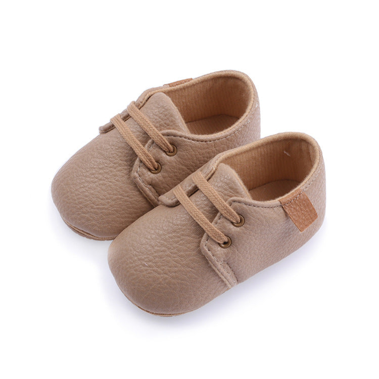 Beige baby shoes with faux leather texture and lace-up design, featuring a small brown fabric tag on the heel. The shoes are placed on a white background.