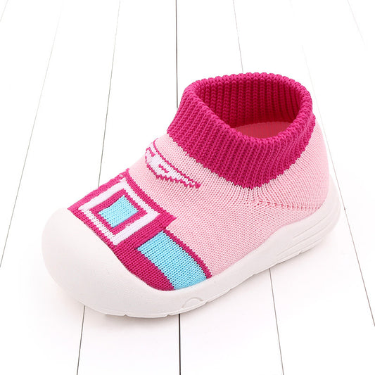 Pink children's sock-style shoe with white sole, featuring a vibrant geometric pattern in blue and dark pink on the top, set against a white, wooden floor background.