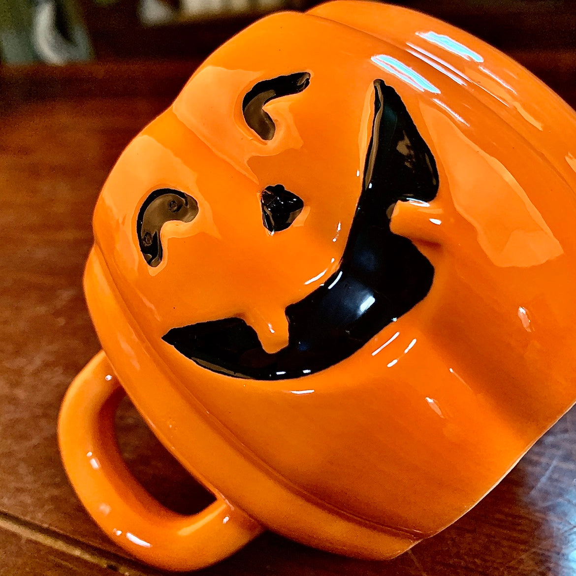 A vibrant orange mug shaped like a jack-o'-lantern with a carved, smiling face, glossy surface, and a curved handle, resting on a wooden table.