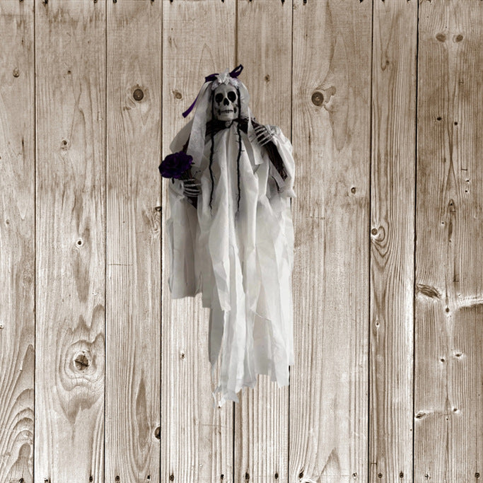 Hanging skeleton dressed in white gauzy fabric, holding a purple flower. The figure has a skull for a face and is adorned with purple ribbons. It is set against a rustic, light wood panel background, providing a contrasting eerie atmosphere.
