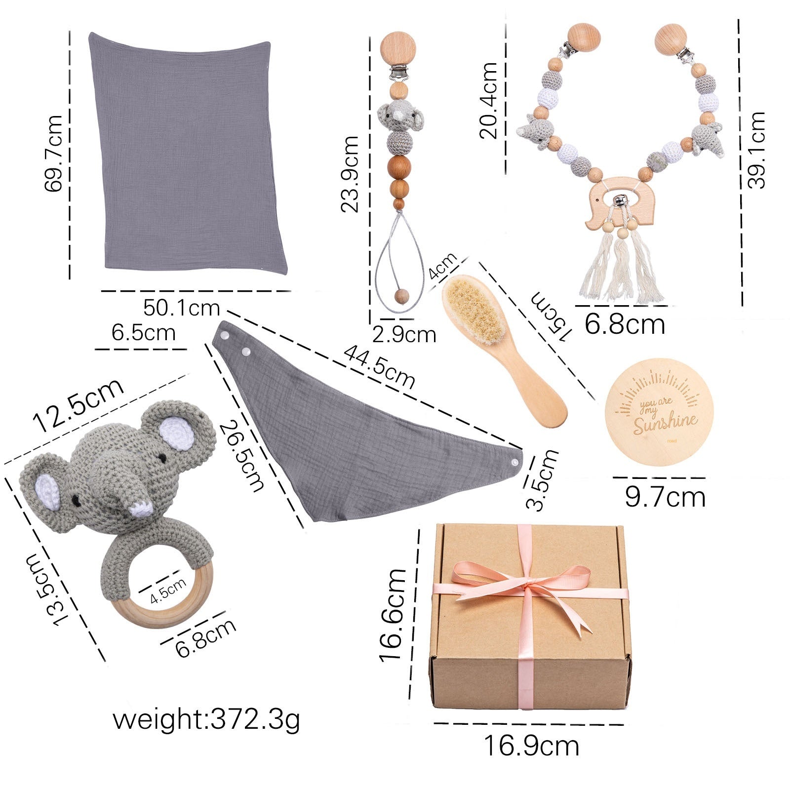 Baby gift set includes a grey blanket, teether, pacifier clip, bandana bib, elephant rattle, wooden brush, engraved plaque, and a gift box tied with a pink ribbon. Measurements are shown.