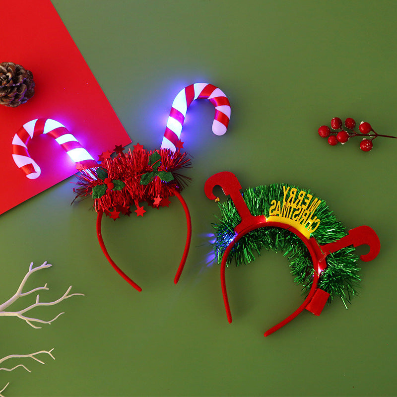 Festive headbands on a green surface: one with glowing candy canes and red tinsel, the other with green tinsel and "Merry Christmas" text. Pine cone and red berries nearby.