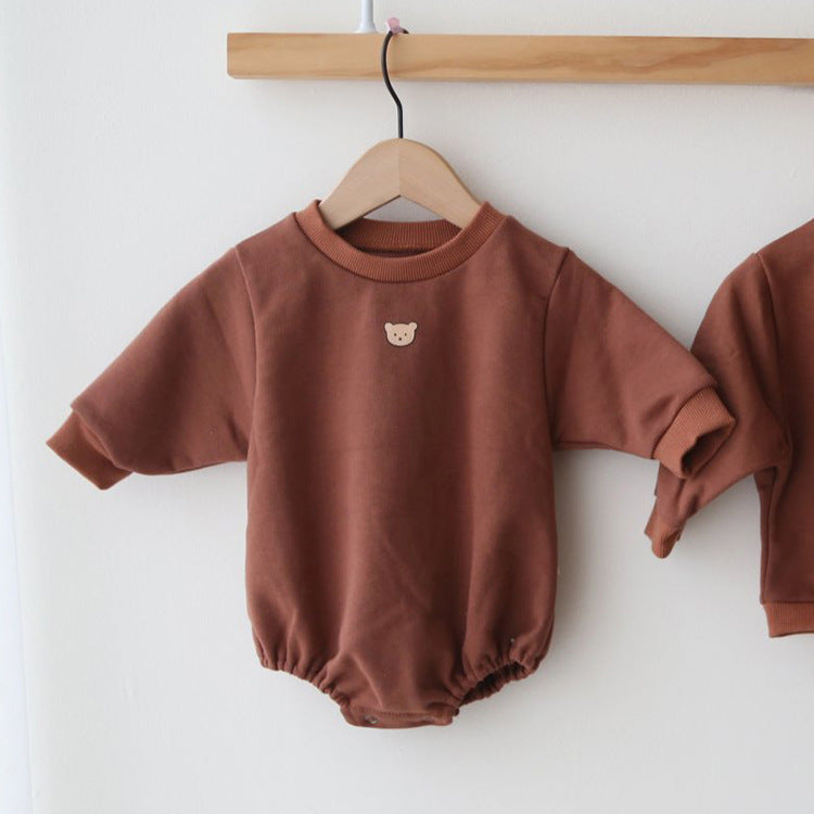 Rust-colored baby romper with long sleeves and a small bear emblem on the chest, hanging on a wooden hanger against a plain white wall.