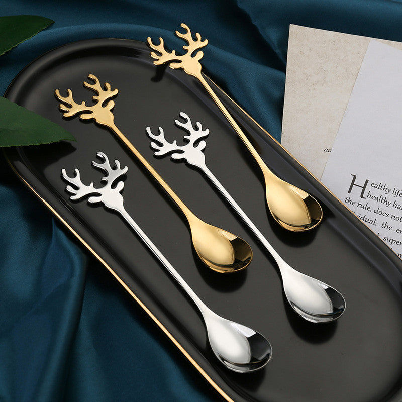 Elegant gold and silver spoons with antler-shaped handles rest on a black tray, placed on teal fabric beside a partially visible page, creating a sophisticated tabletop display.
