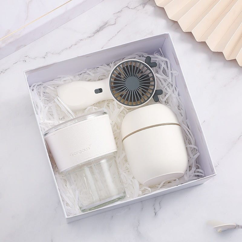 A gift box containing a small white portable fan, a double-walled tumbler with a white band, and an off-white, rounded container, nestled in shredded white paper on a marble surface.
