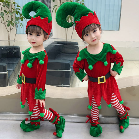 Two young children in matching red and green elf costumes with pointy hats and curly shoes, standing indoors. They wear red and white striped leggings and have festive expressions.