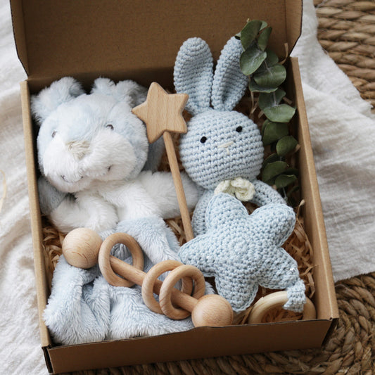 A gift box with a plush bunny, crocheted stuffed rabbit, wooden rattle, star-shaped toy, and eucalyptus leaves, placed on a textured white cloth and woven surface.