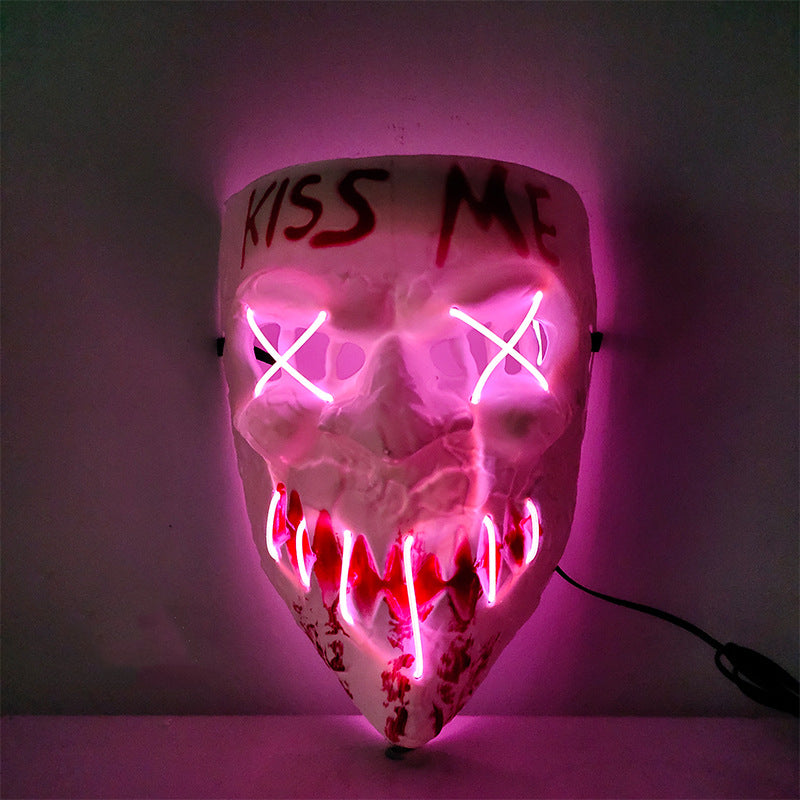 Neon-lit skull mask with "KISS ME" inscribed on the forehead. Eyes are marked with pink crosses and mouth features jagged lines, creating a sinister glow.