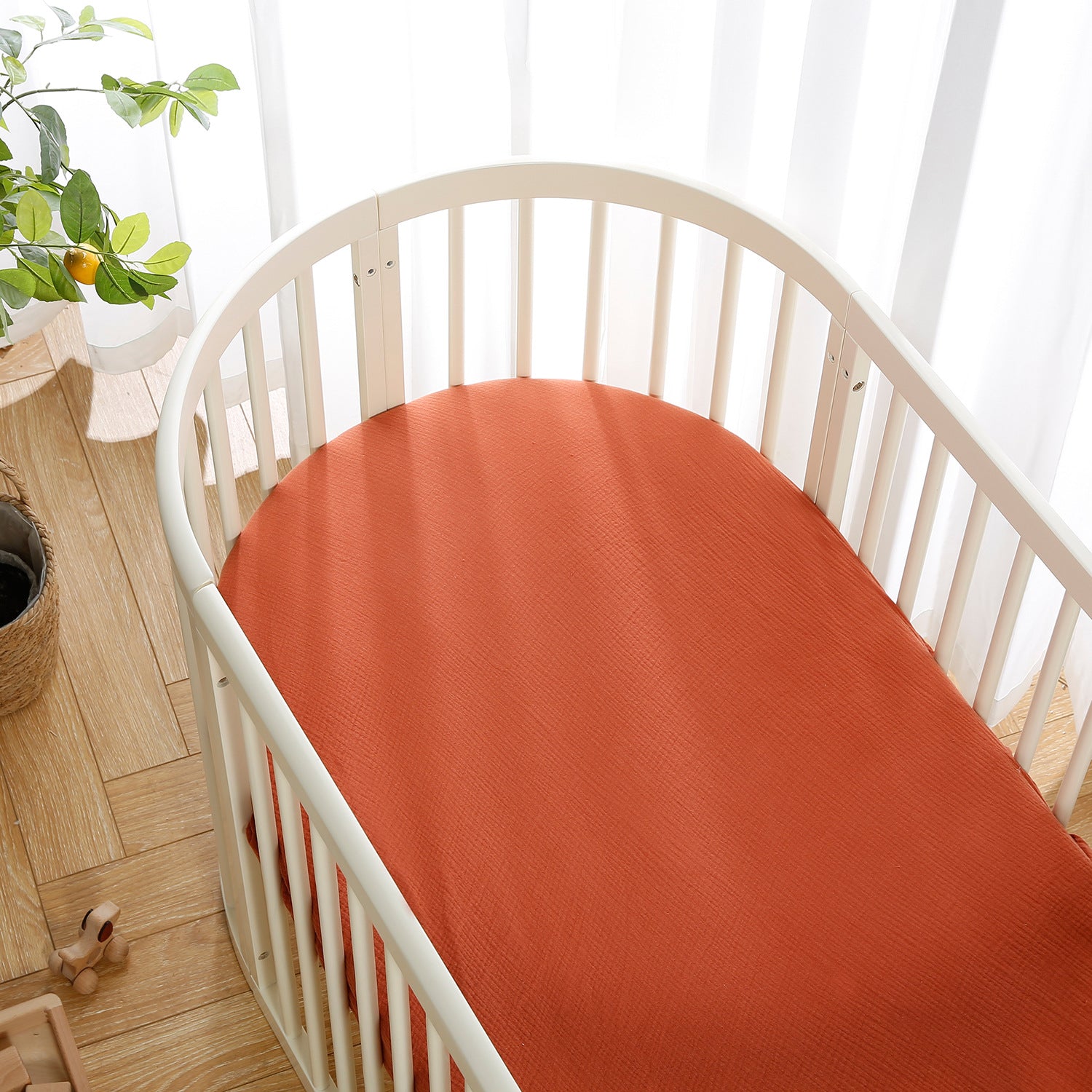 Wooden oval crib with an orange mattress in a bright room, next to a potted plant and wooden toys on a light wood floor. White curtains in the background.