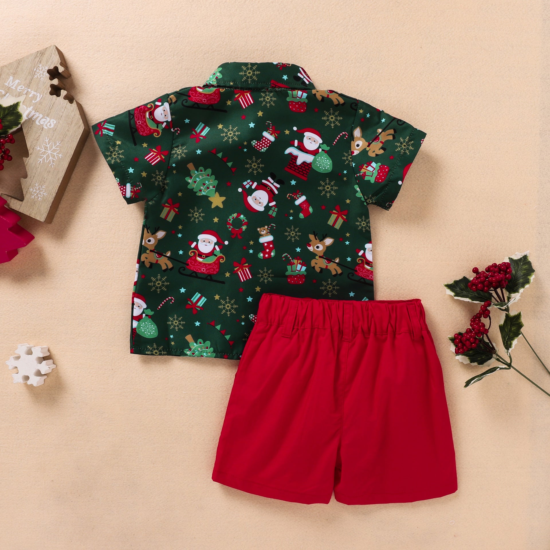 Festive toddler outfit with a green shirt featuring Santa and reindeer, paired with bright red shorts. Decorated background with faux berry sprigs and a wooden Christmas ornament.