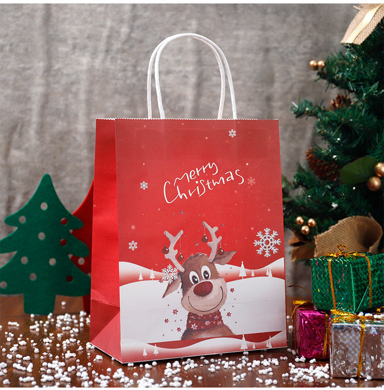 Red gift bag with a cheerful reindeer and "Merry Christmas" text, surrounded by snowflakes. Decorated Christmas tree, gifts, and felt trees on a wooden surface with scattered snow.