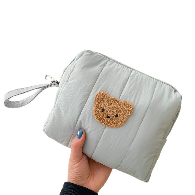 Hand holding a light gray quilted pouch with a strap, featuring a small brown teddy bear face patch. The nails are painted blue.