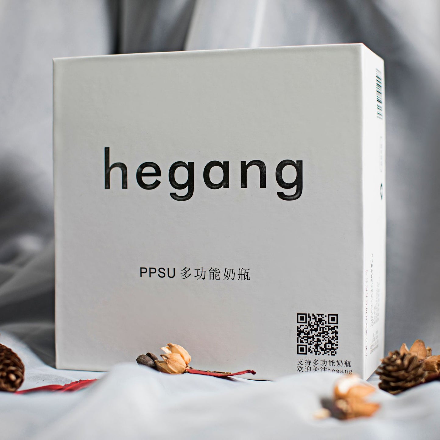 White box with "hegang" text, featuring a QR code and Chinese characters. The box is surrounded by dried flowers and pine cones on a gray fabric background.