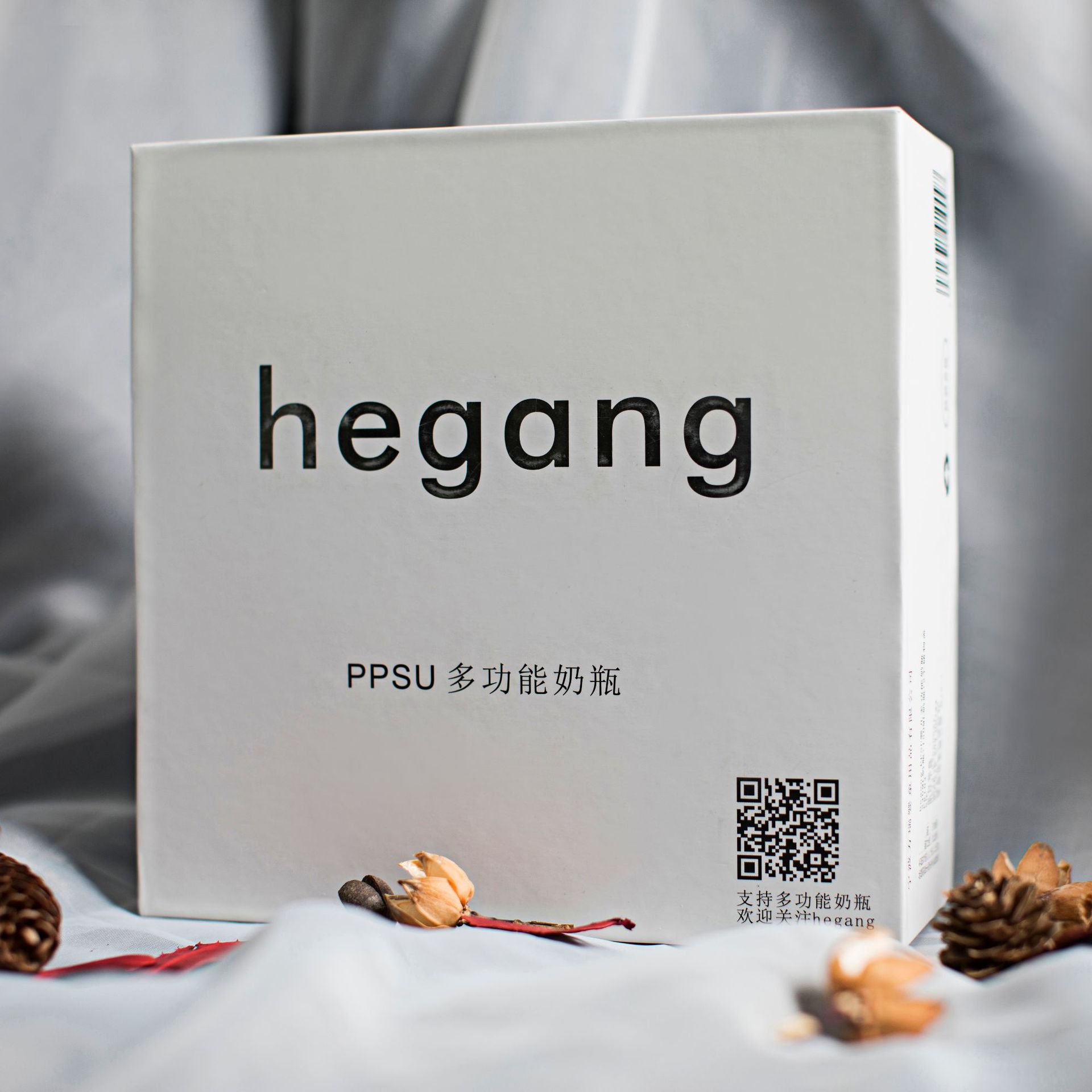 White box with "hegang" text, featuring a QR code and Chinese characters. The box is surrounded by dried flowers and pine cones on a gray fabric background.