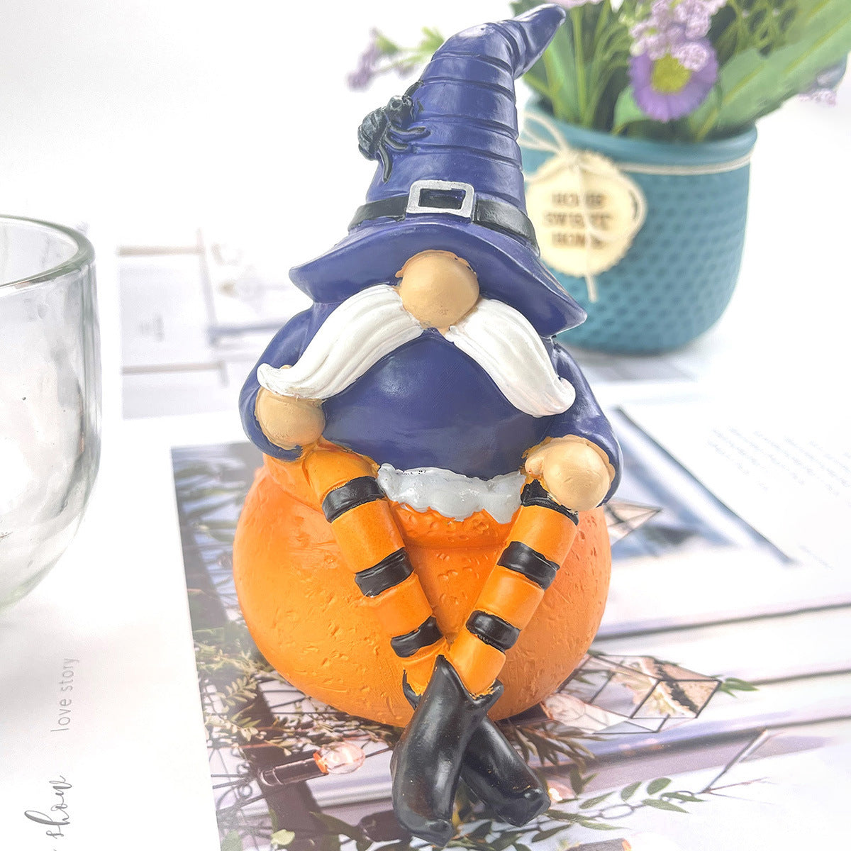 Whimsical Halloween gnome with a white beard, dressed in a purple hat and orange outfit, sits on a pumpkin. In the background, there's a teal pot with purple flowers.