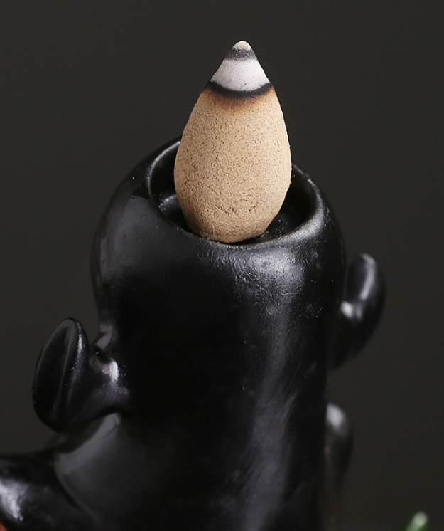 A close-up of a black ceramic incense holder with a tan cone incense placed inside. The holder has a glossy finish and is designed with small ear-like shapes on its sides.