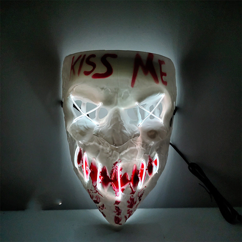 White skull mask with glowing white stitched eyes and red-lit jagged mouth, features "KISS ME" written in red on the forehead. The mask is mounted on a dark wall.