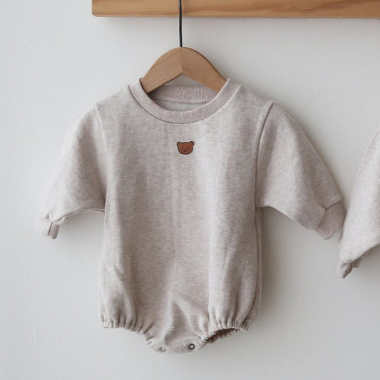 Cream-colored baby onesie with short sleeves and a small brown bear face on the chest, hanging on a wooden hanger against a white background.