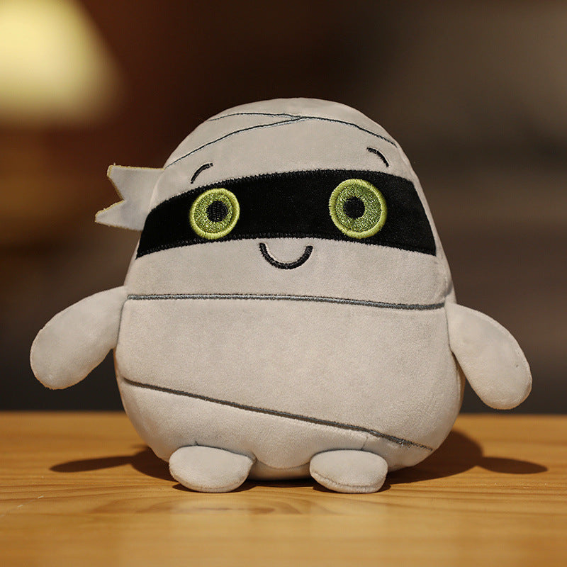 Cute plush toy resembling a mummy with a black mask, large green eyes, and a smiling face, seated on a wooden surface with a blurred background.