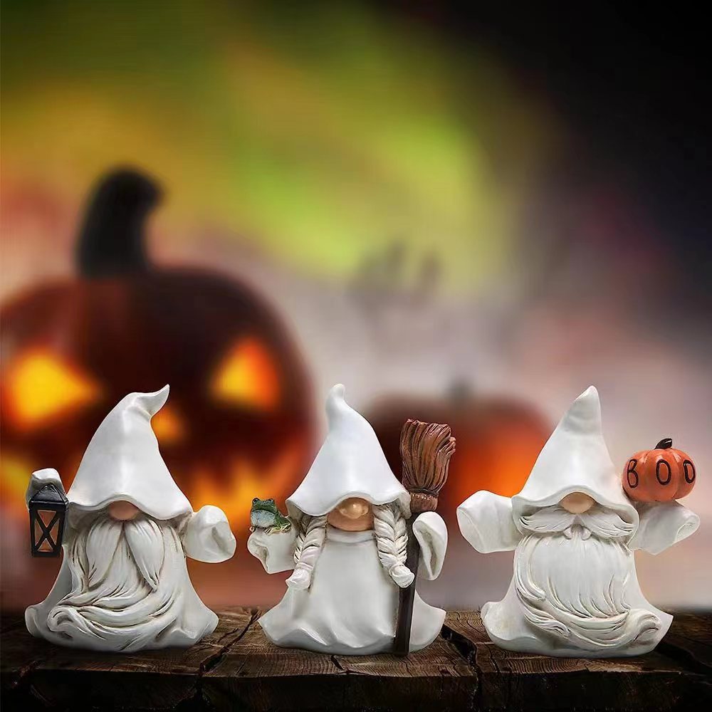 Three ghostly gnome figurines in white cloaks on a wooden surface. One holds a lantern, another a frog and broom, and the third a pumpkin labeled "Boo." Jack-o'-lanterns glow in the background.