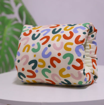 A colorful, folded cloth diaper with a playful abstract pattern rests on a white surface. The background features a blurred green leaf design, adding a touch of natural decor.