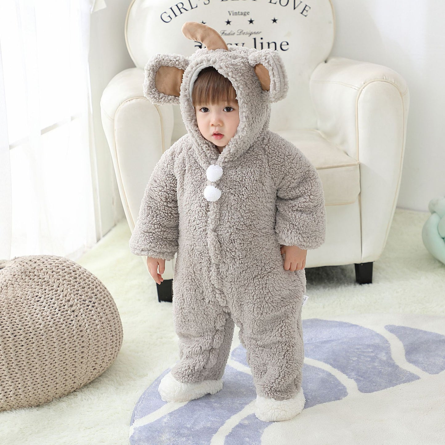 A toddler in a fluffy koala costume with large ears and pom-pom buttons stands on a patterned rug, in a cozy room with a white armchair and soft lighting.