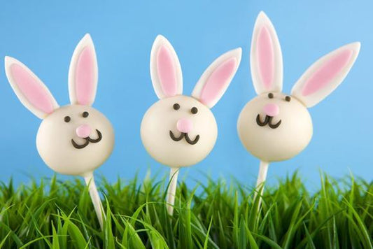 Three bunny-shaped cake pops with pink ears and noses, and cute faces, stand upright among vibrant green grass against a bright blue sky background.
