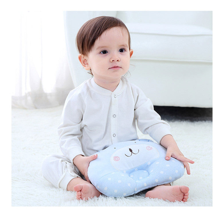 A toddler in white pajamas sits on a soft white rug holding a blue polka-dotted cushion with a cute bear face. The background features a white sofa and sheer curtains.