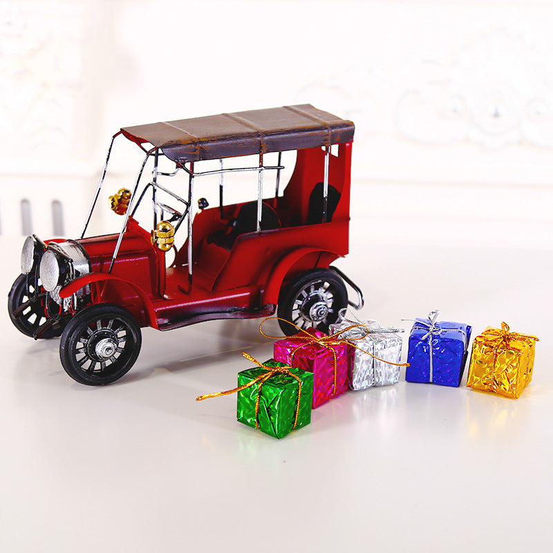 A vintage red toy car with a black roof sits on a white surface, surrounded by five small, colorful gift boxes wrapped in metallic paper: green, pink, silver, blue, and gold.