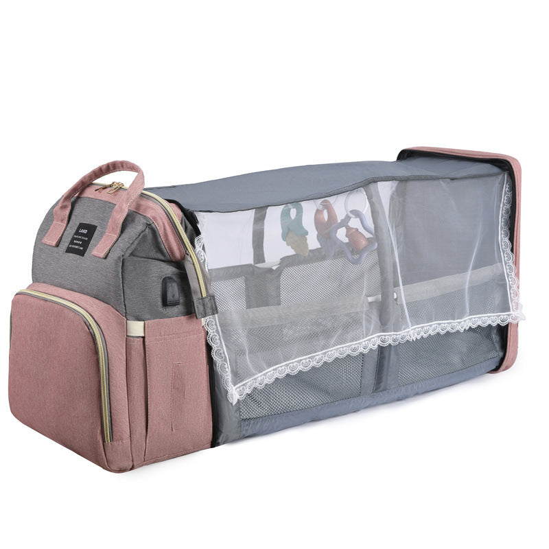 Gray and pink multifunctional baby bag with side storage, front pocket, and mesh-lined fold-out crib extension featuring hanging toys, designed for convenience and baby comfort.