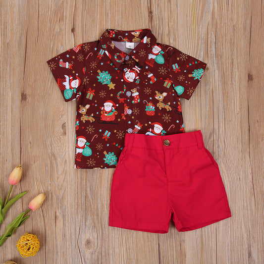 Children's festive outfit with a short-sleeved shirt featuring a Christmas-themed pattern and bright red shorts, laid out on a wooden floor alongside tulips and a decorative ball.