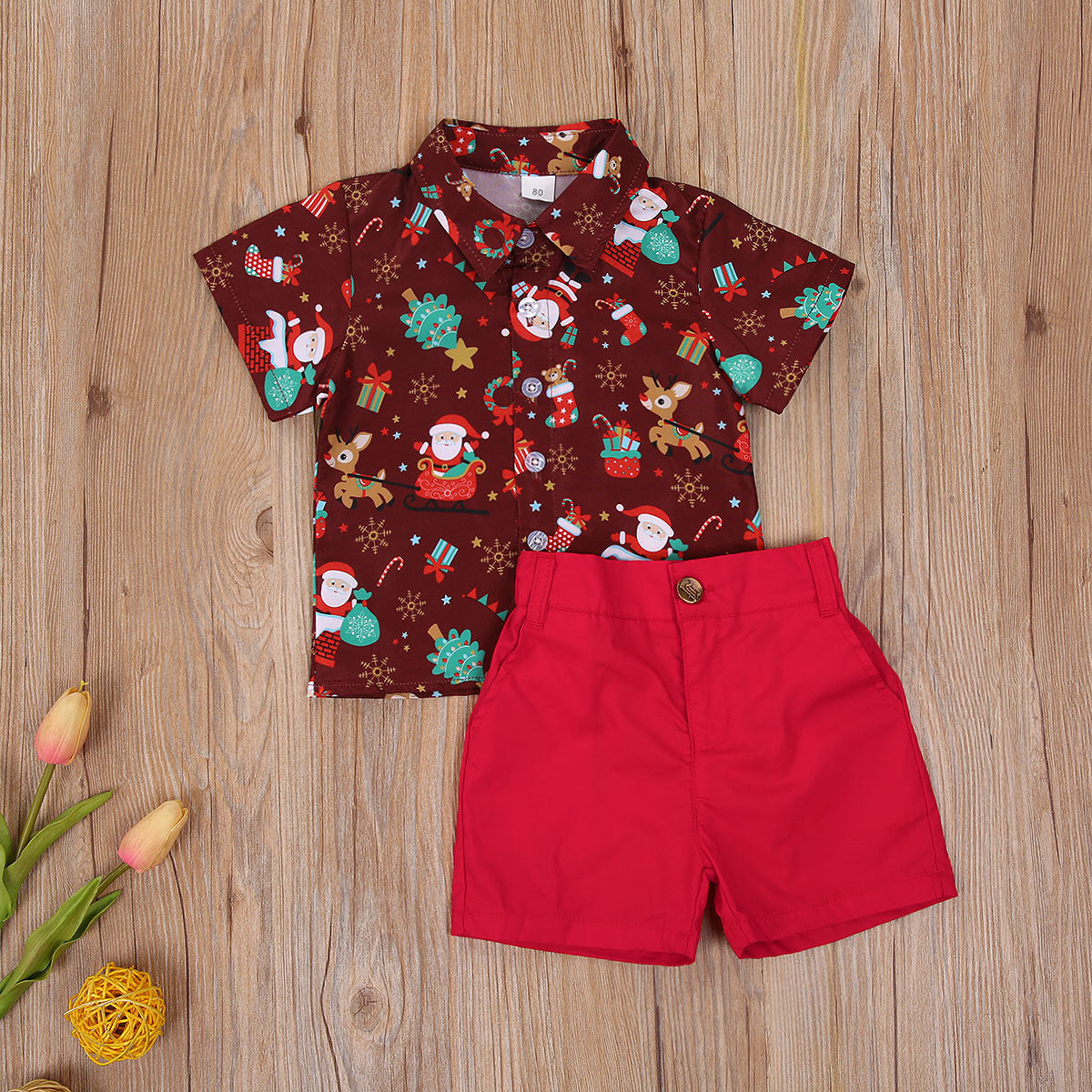 Children's outfit on wooden floor: a festive shirt with Santa and reindeer patterns, paired with red shorts. Nearby, yellow yarn and pink tulips add a playful touch.