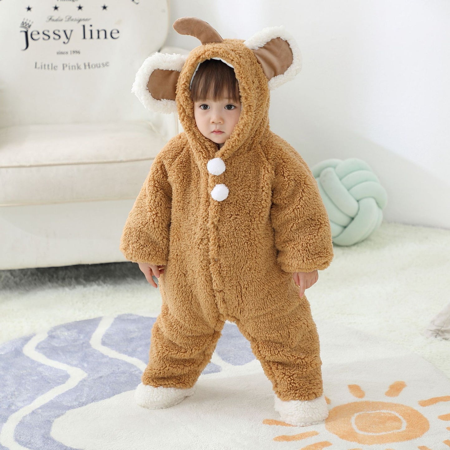 A toddler in a fuzzy brown deer costume with antlers and white accents stands on a soft rug, next to a couch and pastel knot cushion in a bright room.