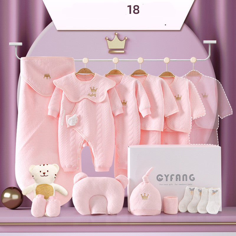 A pastel pink baby clothing set including onesies, hats, mittens, and socks, featuring small crown designs. A teddy bear and a branded box are also displayed in the scene.