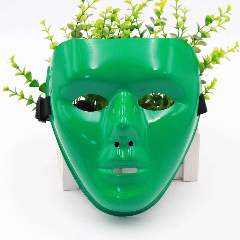 A shiny green mask with eye slits and a small vent, set against a background of leafy plants, creating a whimsical effect as though the plants are sprouting from the mask.