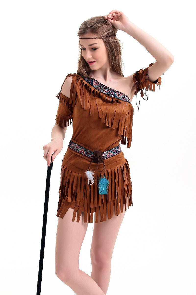 Woman in a fringed brown costume dress with colorful patterns and feathers, looking down. She holds a stick and adjusts her hair, set against a plain white background.