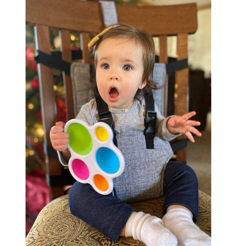 A baby sitting in a high chair, wearing a gray outfit and holding a colorful toy with circular pads. The baby looks surprised, with an open mouth and wide eyes.