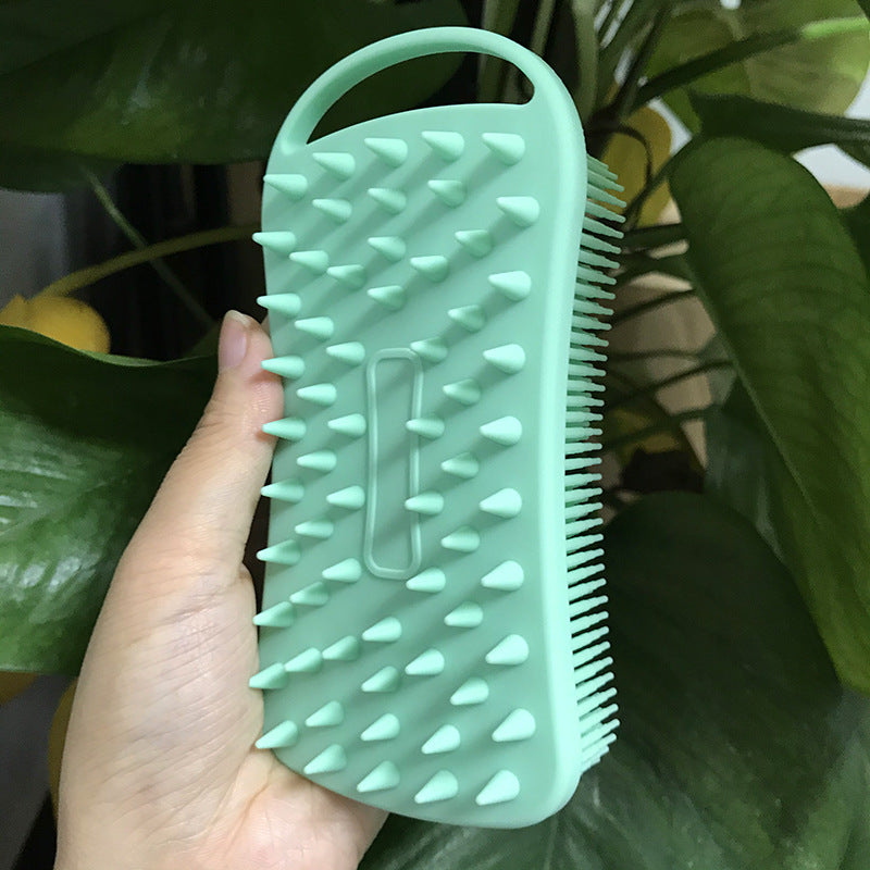 A hand holding a green silicone scrubber with short spikes on one side and long bristles on the other, set against a backdrop of green leaves.
