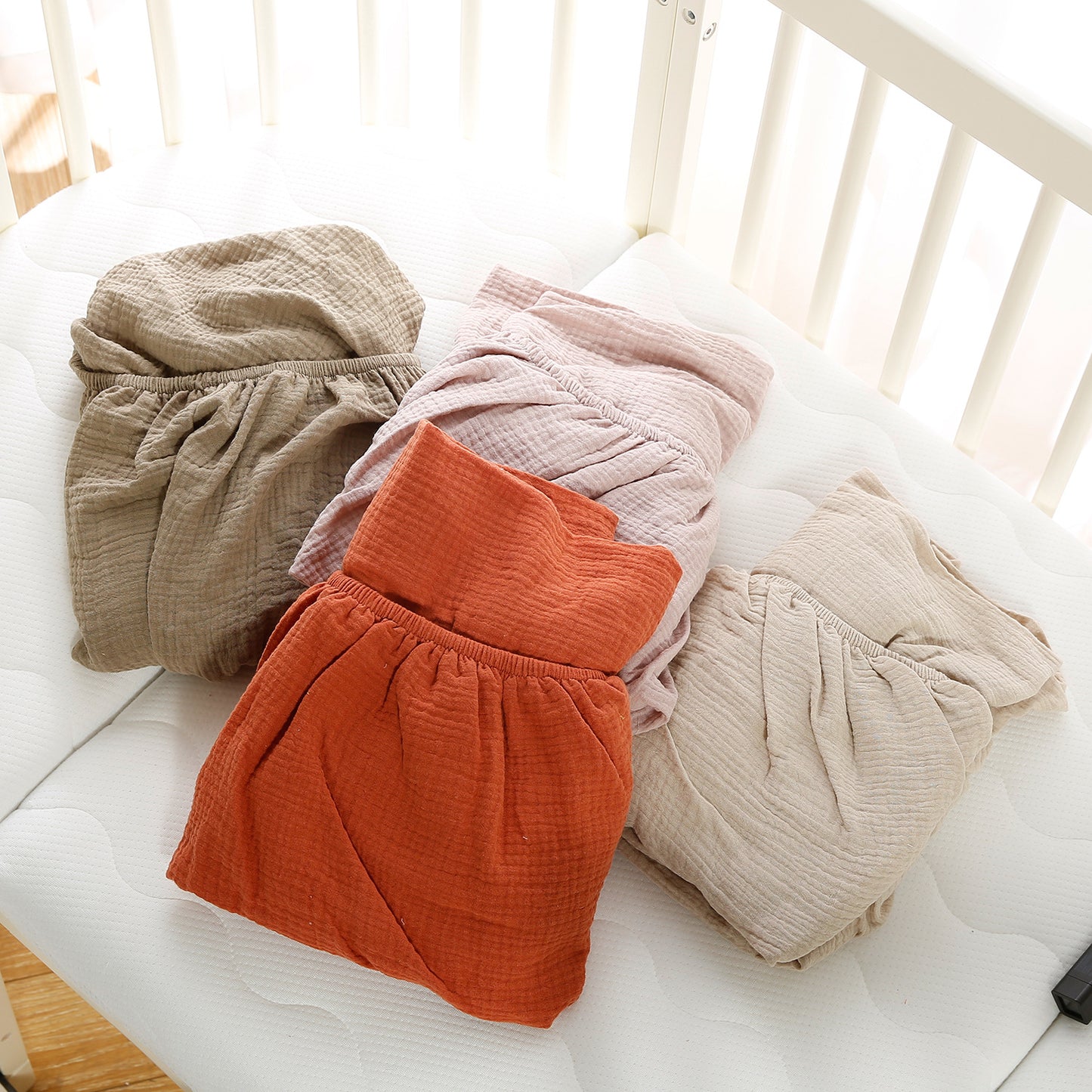 Four pairs of crinkled fabric pants in beige, pink, orange, and taupe are neatly folded on a white mattress by the side of a crib, showcasing a soft and comfortable texture.