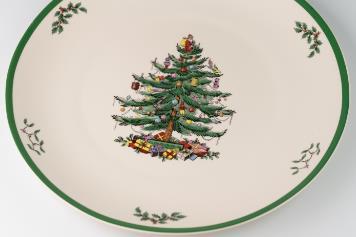 A decorative plate with a green-rimmed edge, featuring a Christmas tree adorned with ornaments and gifts underneath. Small holly sprigs accent the plate's border.