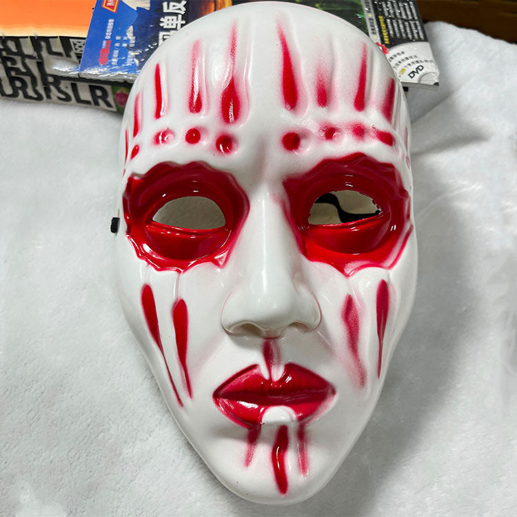 White mask with red markings highlighting eyes, mouth, and vertical lines across the face, set against a soft background with partially visible books.