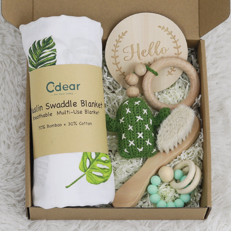 Baby gift set in a box featuring a muslin swaddle blanket with leaf print, a wooden "Hello" sign, a green cactus teether, baby brush, and a mint bead bracelet.