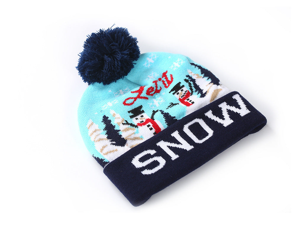 Light blue beanie with dark blue pom-pom, featuring a snow scene with snowmen and trees. "Let it Snow" is written in red and white across the hat and cuff.